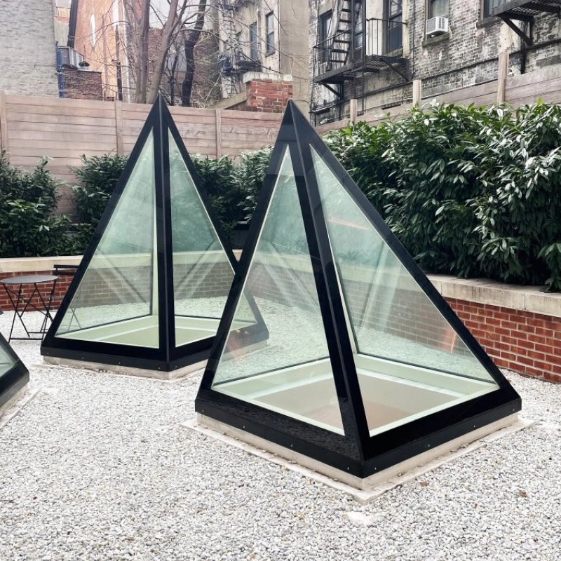 Two modern glass and black-frame pyramidal structures are placed on a gravel surface in a courtyard, surrounded by low greenery and brick walls. An apartment building with fire escapes is visible in the background.