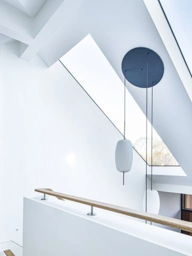 Bright, modern hallway featuring a white exterior, large skylights, and a wooden railing. The minimalist design includes angular architectural elements and a round pendant light.