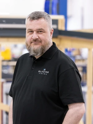 A man with a beard poses confidently in a black polo shirt, standing in a workshop filled with wooden frames and machinery.
