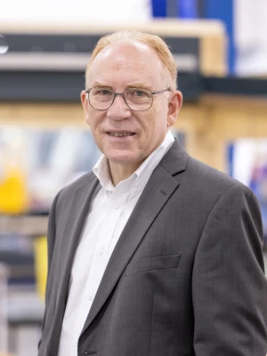 A man with glasses stands confidently in a workshop, wearing a dark suit and white shirt. He has short, light brown hair and is smiling slightly while looking at the camera. Behind him is a blurred background of wooden structures and work equipment.