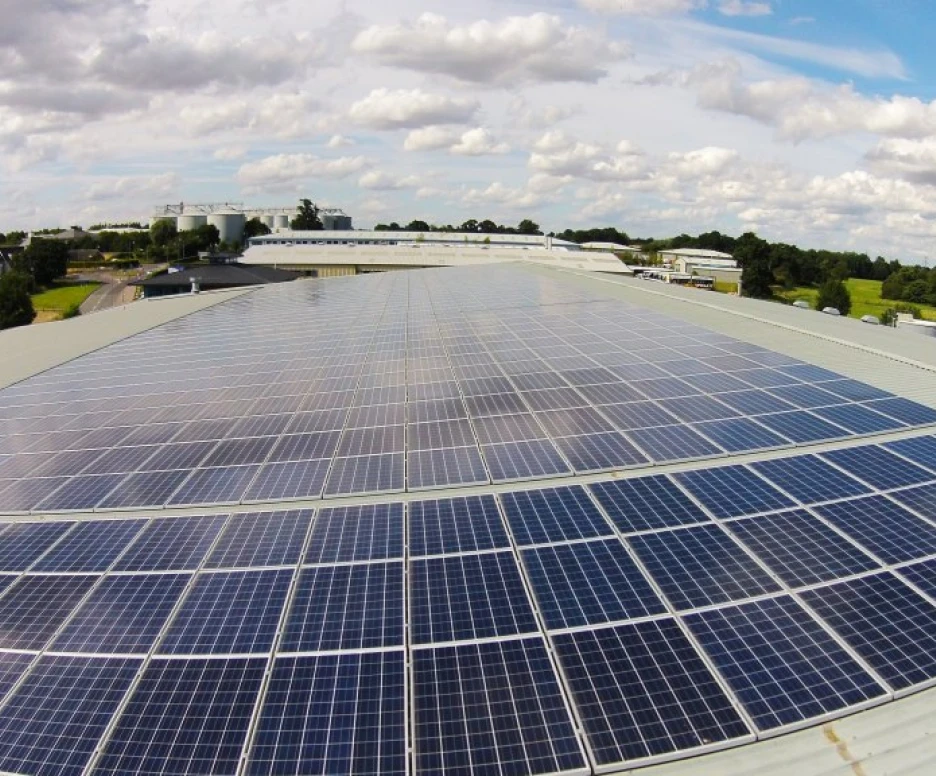A rooftop covered with numerous solar panels under a partly cloudy sky. In the background, a landscape features green fields and buildings.