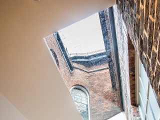 A modern interior space featuring a skylight that illuminates the room with natural light, revealing rain on the glass. Exposed brick walls contrast with smooth white surfaces, creating a contemporary aesthetic.