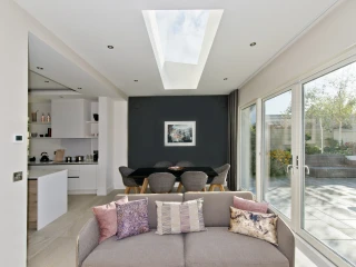 A modern interior showcasing an open living space with a couch adorned with decorative pillows. In the background, a dining area features a black table surrounded by gray chairs, and large glass doors lead to a garden. A skylight above provides natural light, illuminating the room.