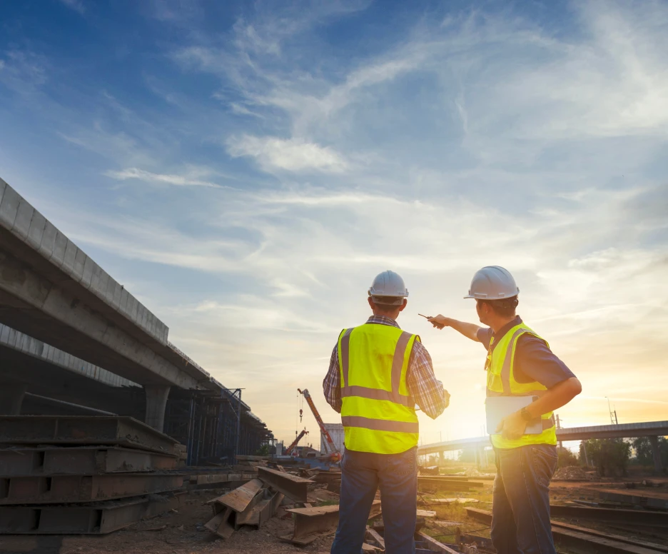 Two construction workers in yellow vests and hard hats stand on a construction site, discussing plans against a backdrop of a vibrant sunset and partly cloudy sky.