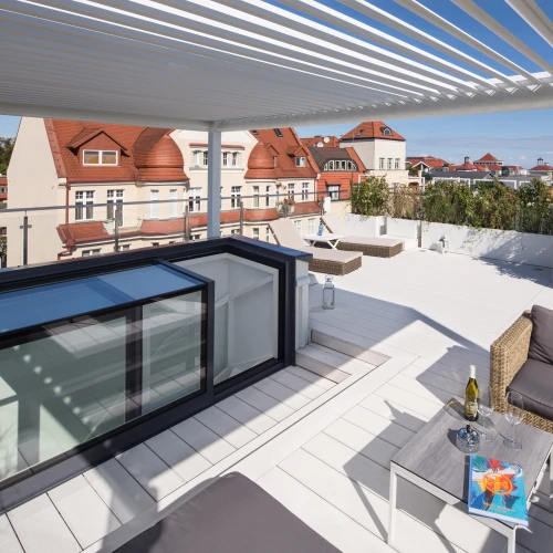 A spacious rooftop terrace featuring a modern seating area with a gray sofa and a coffee table. Sunlight filters through white slatted pergola shadows. In the background, red-roofed buildings and a clear blue sky complete the scene.