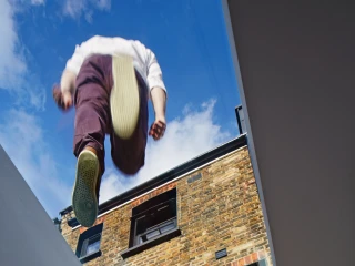 A person is jumping above an open space in a modern building, with a view of a brick wall and blue sky visible through the skylight. Shadows are cast on the interior surface below.