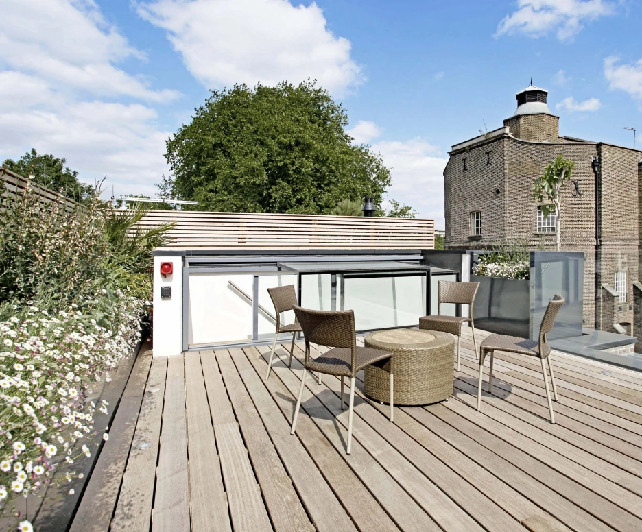 A modern rooftop terrace featuring a wooden deck, a round table, and four chairs. Lush green trees and a brick building are visible in the background under a cloudy blue sky.