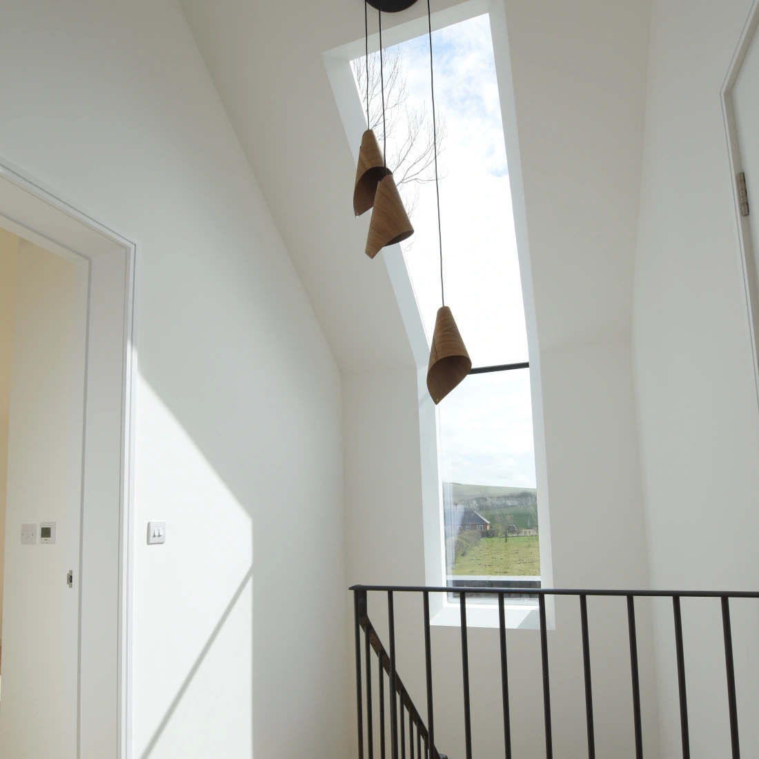 A modern hallway with a sloped ceiling features three pendant lights hanging from a skylight. A black railing borders the wooden staircase, and sunlight streams through the window, casting shadows on the floor.