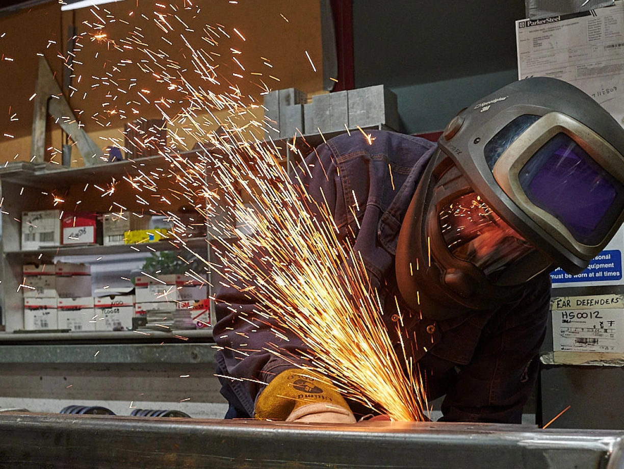 A welder in a protective helmet works on metal, creating bright sparks that illuminate the workshop. Tools and materials are visible in the background, indicating an industrial setting.
