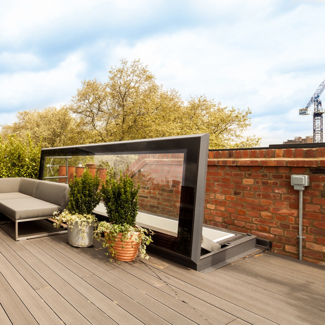 A modern rooftop terrace with gray sectional seating, a small round table, and potted plants. Surrounding greenery adds a natural touch, while a glass windbreak offers a stylish barrier. A blue sky with scattered clouds is visible overhead.