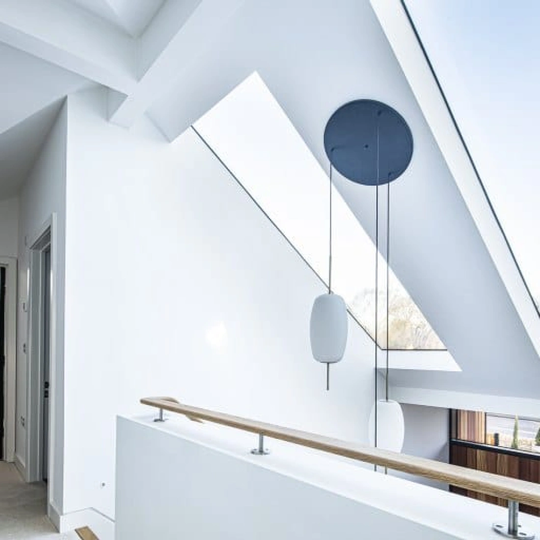 Bright, modern hallway featuring a white exterior, large skylights, and a wooden railing. The minimalist design includes angular architectural elements and a round pendant light.